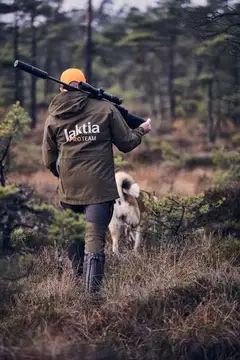 A hunter wearing a Jaktia Pro Team jacket walking through woodland with a dog.